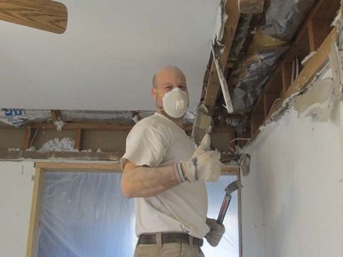 Worker wearing a mask and gloves repairs ceiling drywall with a hammer in a room under renovation