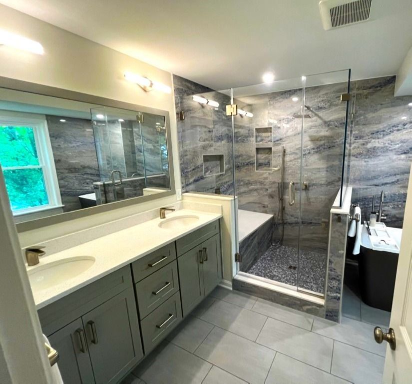 Modern bathroom with double vanity, large mirror, glass shower, gray tile, and bathtub with stone-look walls