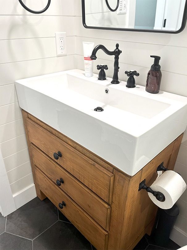 Wood vanity with white sink and black faucet in a tiled bathroom, with toilet paper holder on the side