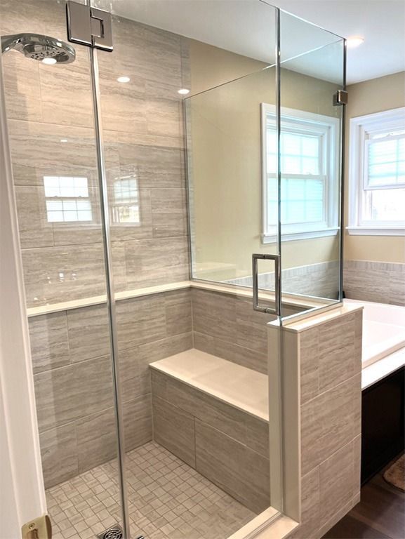 Modern glass shower with built-in bench, gray tile, and beige stone surround under bright window light