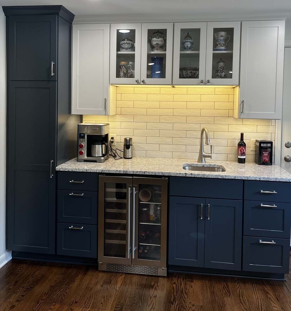 Modern kitchen with navy and white cabinets, lit tile backsplash, marble countertop, and wine fridge