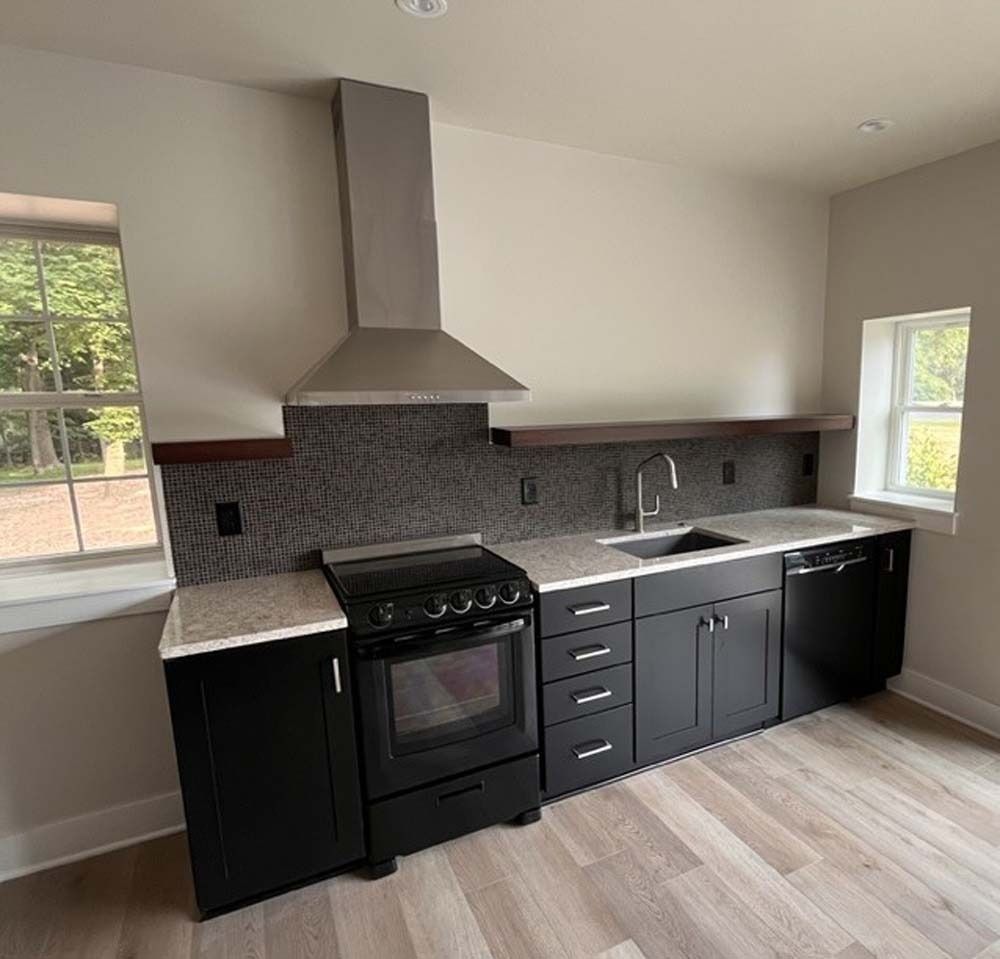 Modern kitchen with black cabinets, gray tile backsplash, stainless range hood, and light wood floors