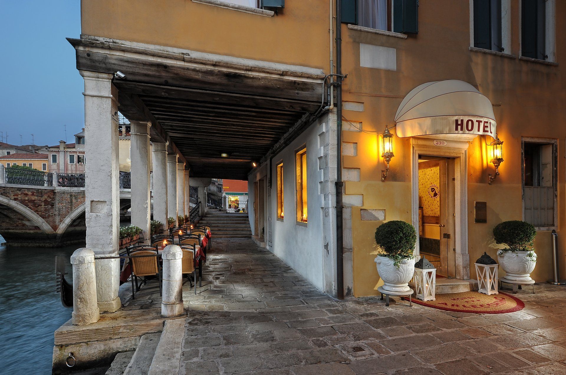 Covered walkway beside a lit hotel entrance by a canal at dusk, with umbrellas and potted topiary trees