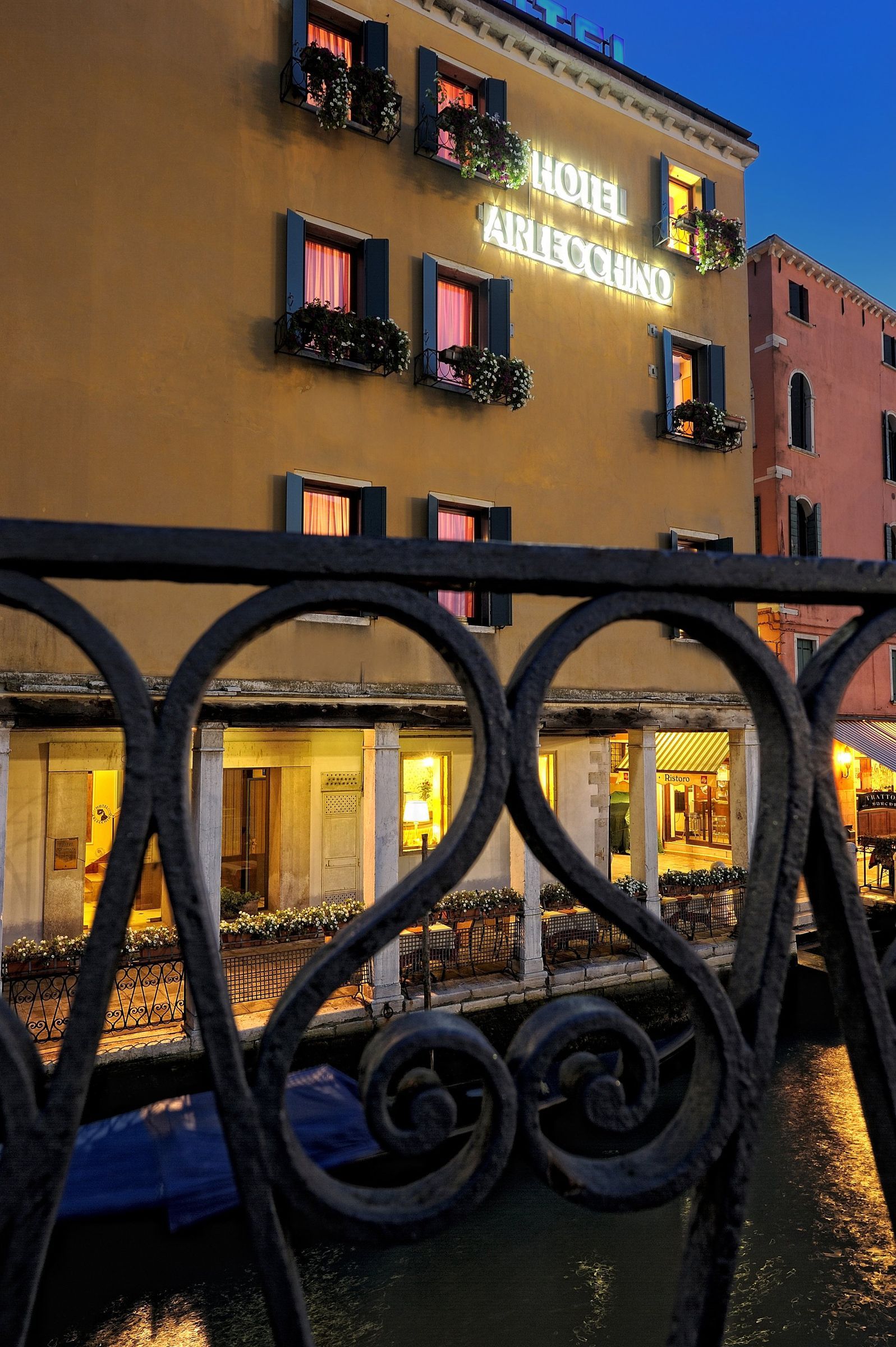 Canal-side hotel at dusk with lit windows and black wrought-iron bridge railing in the foreground