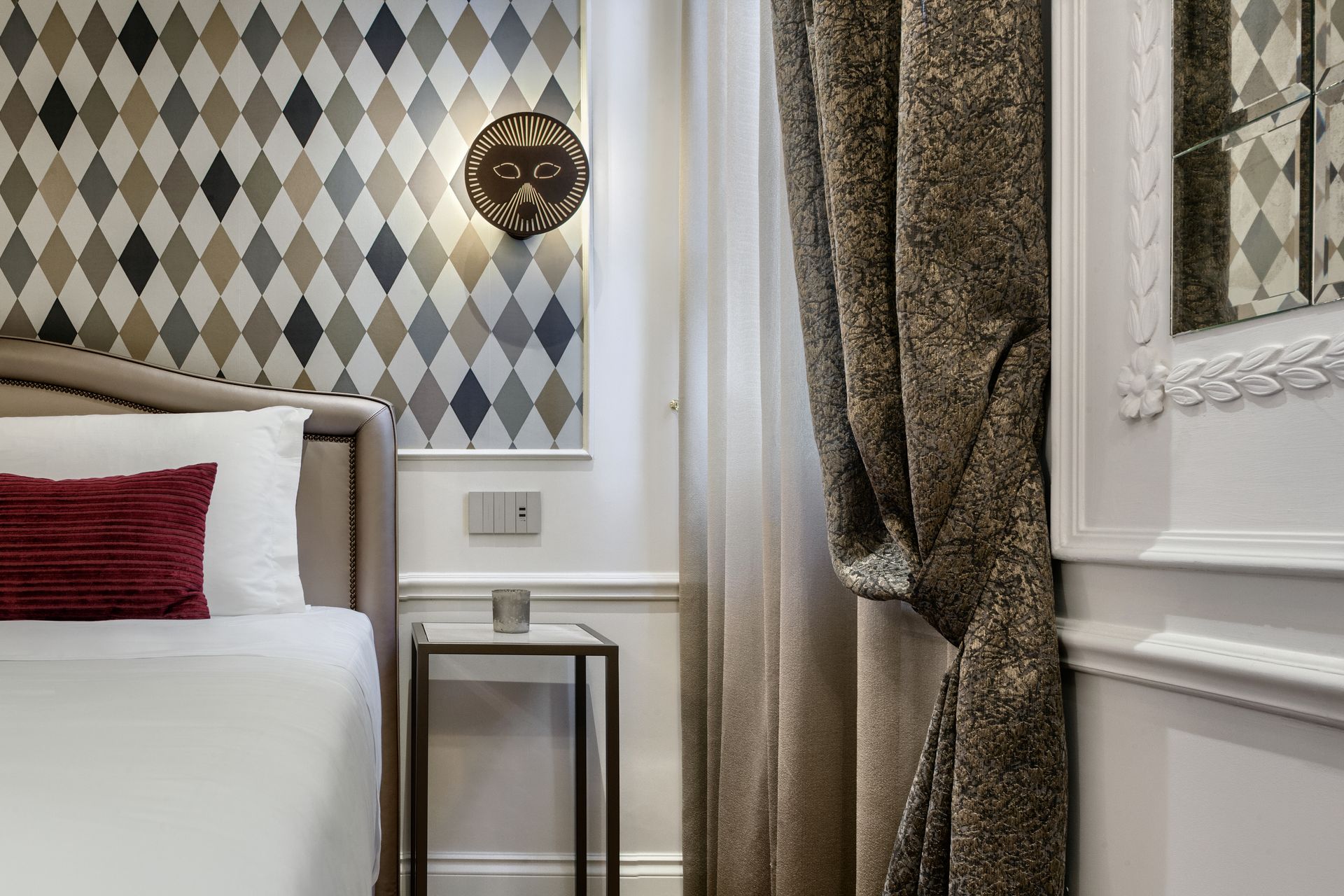 Hotel room corner with patterned wallpaper, bed edge, side table, and heavy curtains