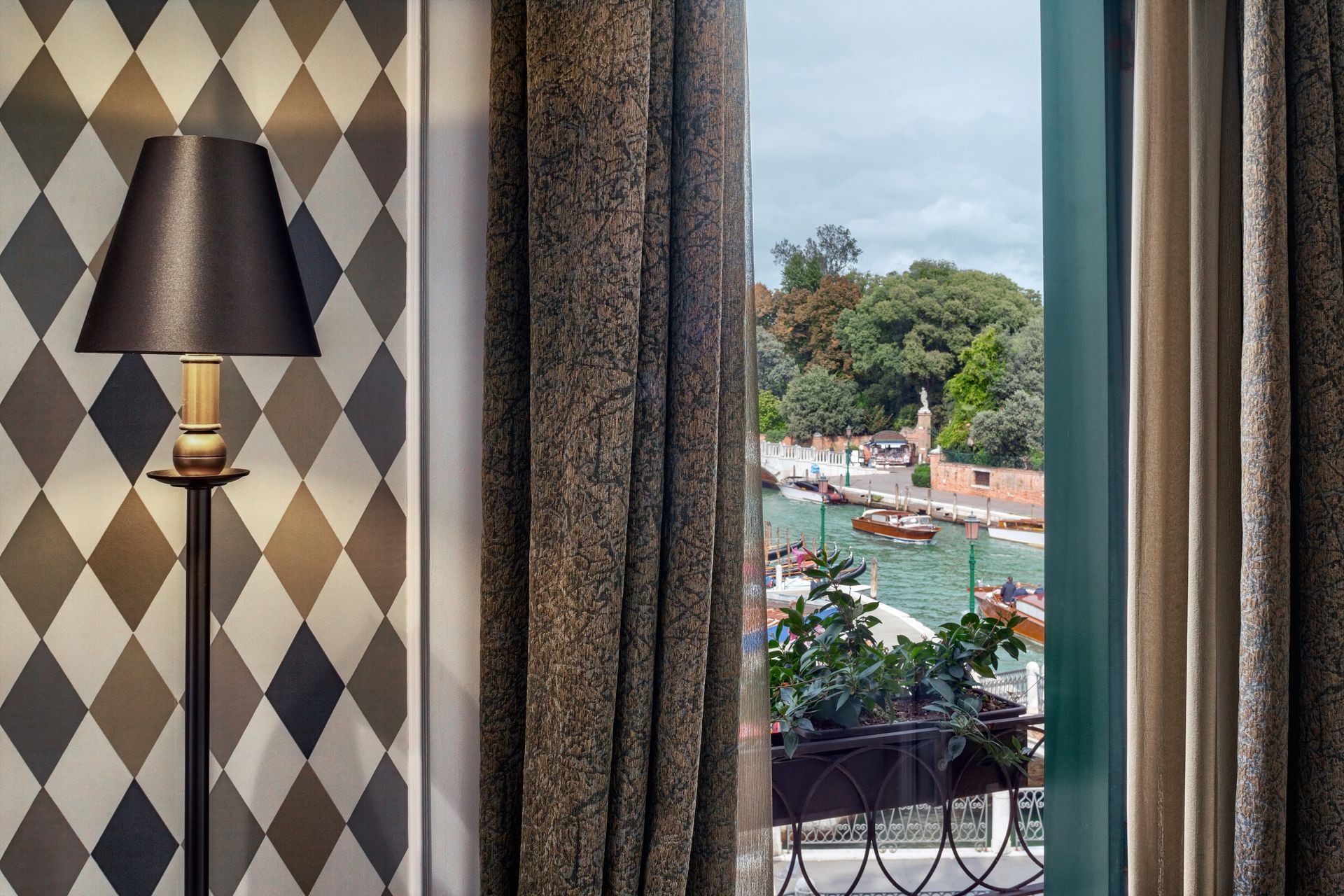 View through a hotel window of a river with boats, framed by curtains and a patterned wall lamp inside.