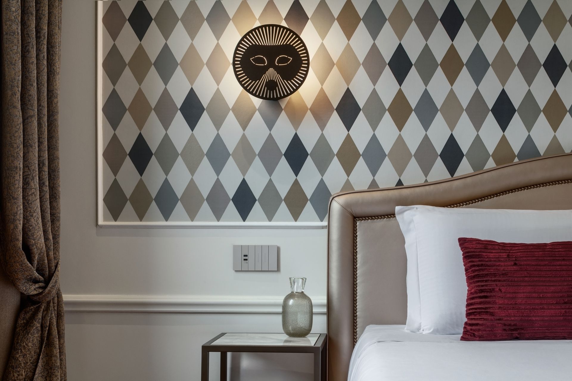 Bedroom corner with patterned wall art, bedside table, and bed with white bedding and red pillow.