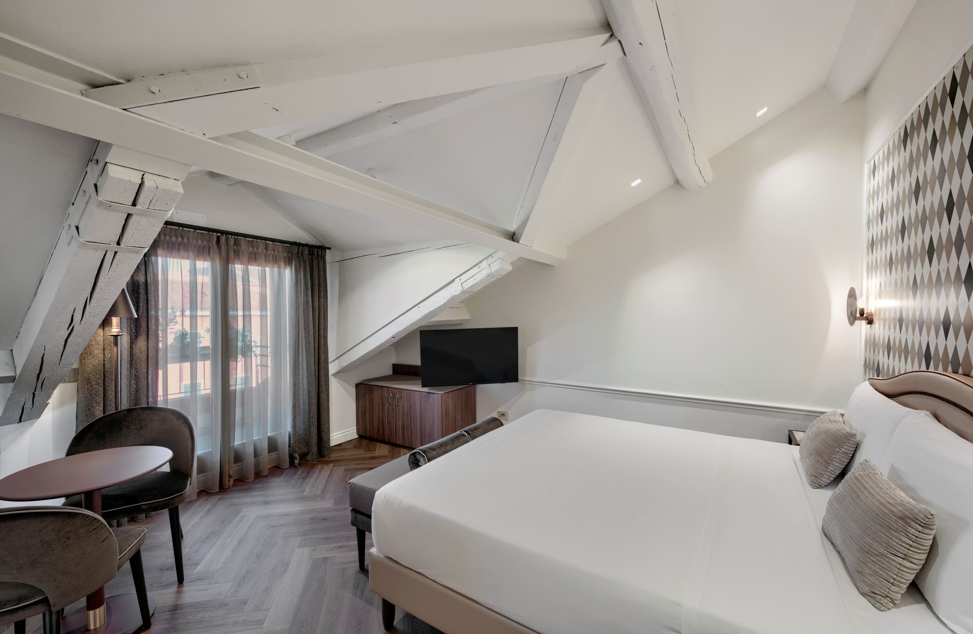 Modern hotel room with white bed, sloped ceiling, desk, and balcony doors with sheer curtains