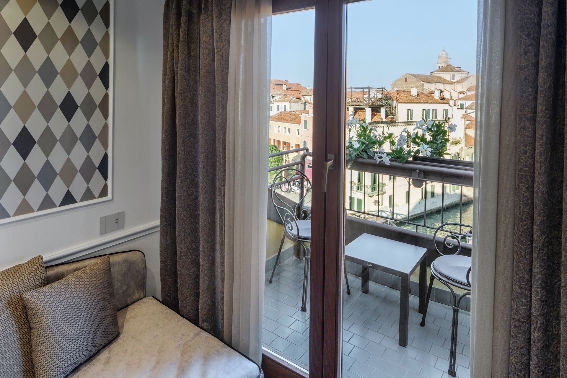 Cozy balcony view through glass doors with chairs, table, and a sunlit cityscape beyond