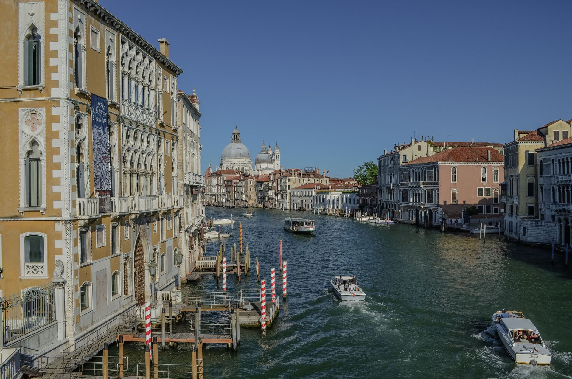 Canale a Venezia con barche, edifici storici e una chiesa con cupola in lontananza, sotto un cielo azzurro e limpido