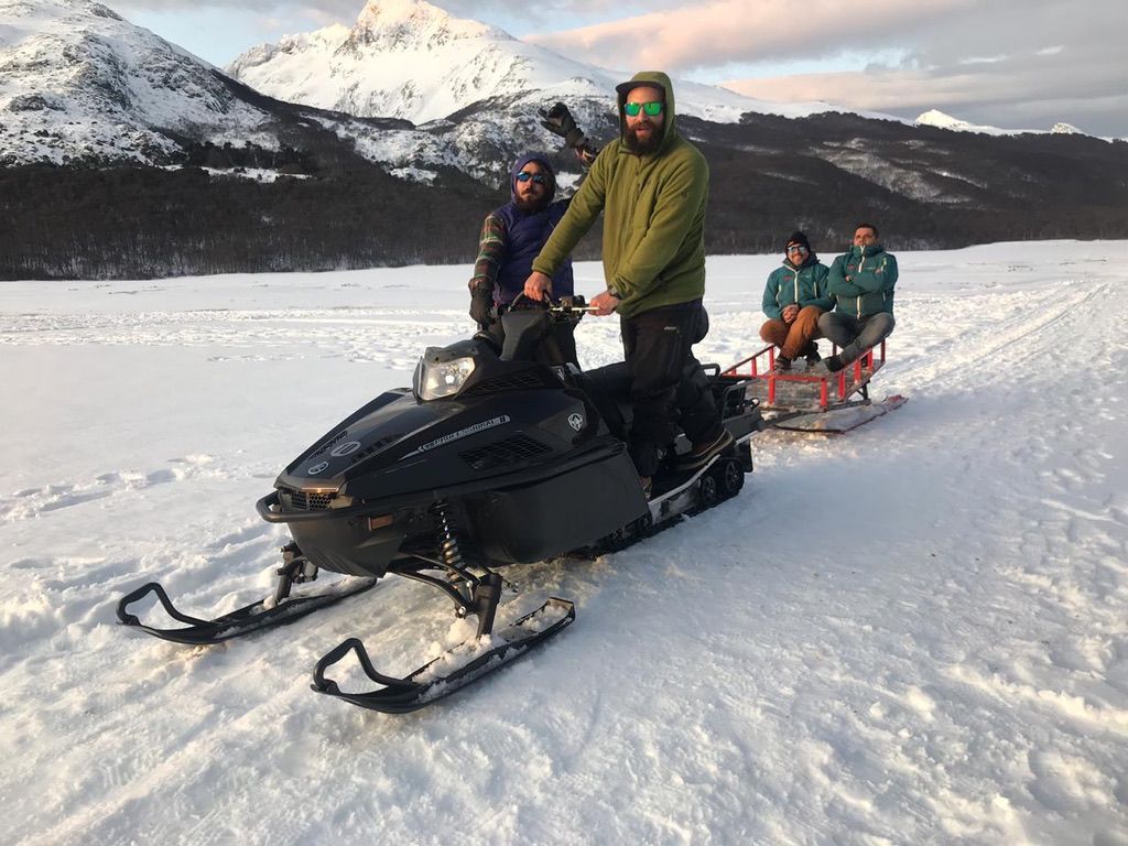 Moto de nieve tirando de un trineo a través de un paisaje cubierto de nieve con montañas al fondo, transportando a cuatro personas.