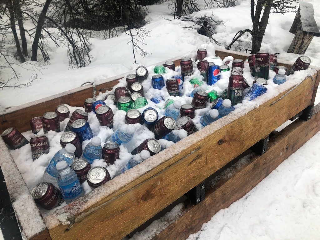 Nevera portátil de madera llena de nieve, latas y botellas en un entorno nevado al aire libre.