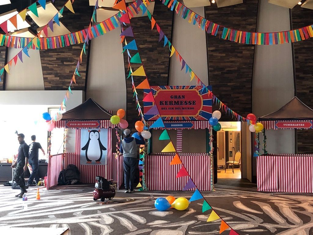 Evento de temática circense en interiores con banderines y puestos de colores. Los trabajadores están montando la decoración.
