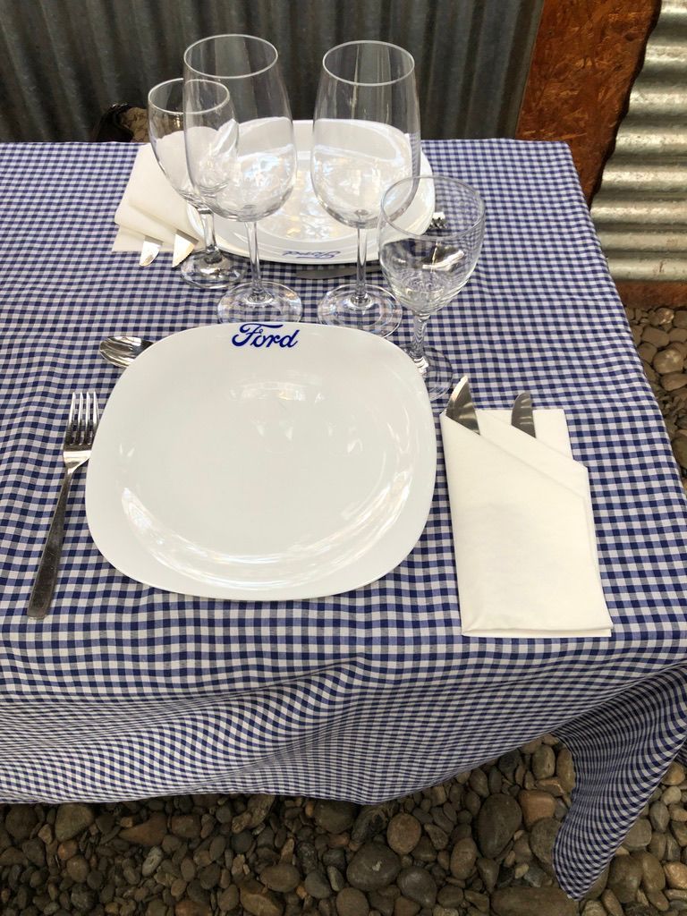 Mesa con el logotipo de Ford en el plato, con copas de vino, servilletas y cubiertos sobre un mantel a cuadros.