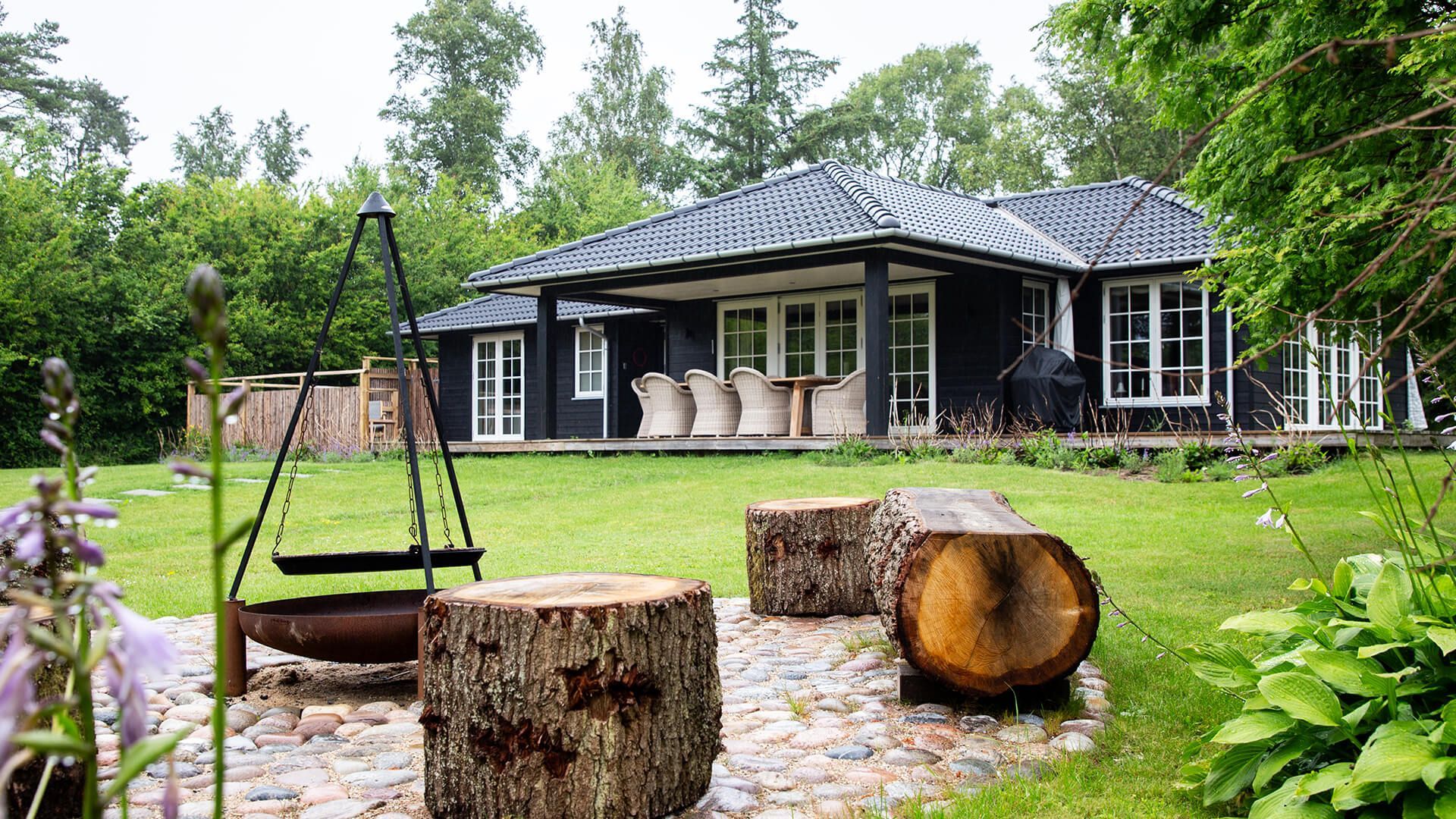 Baghave med sort hus, terrassesæder, bålplads med bålstole og trebenet grill ved en grøn græsplæne