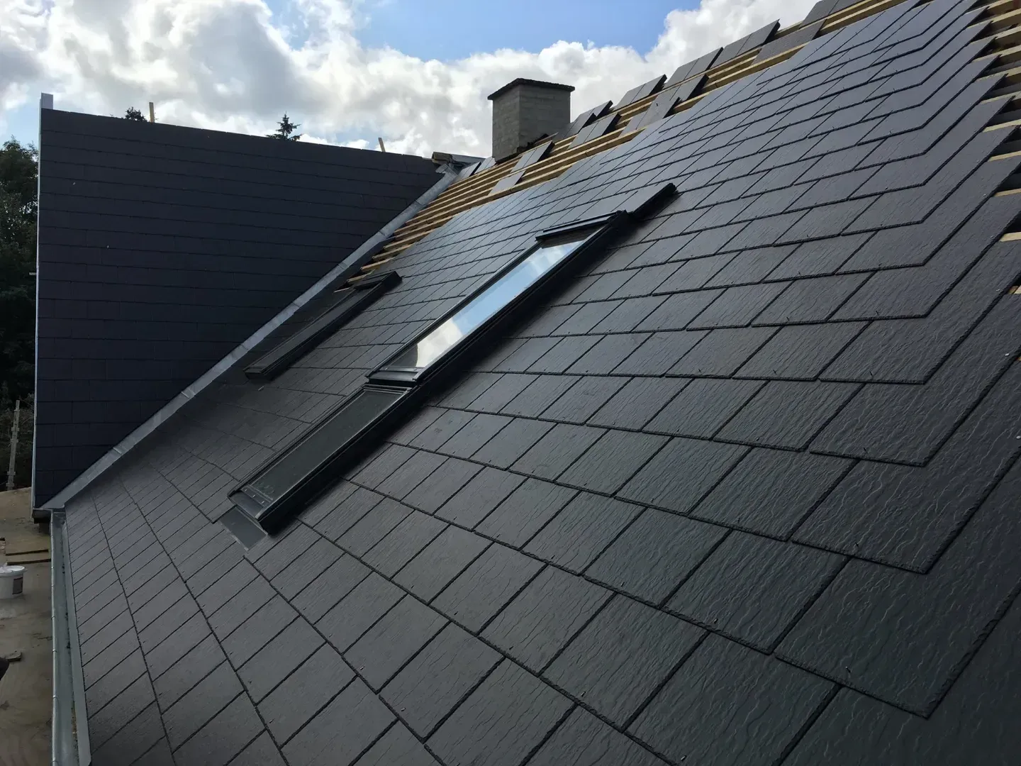 Træramme af et hus under opførelse, blå himmel, byggematerialer og et delvist færdiggjort hus i baggrunden.