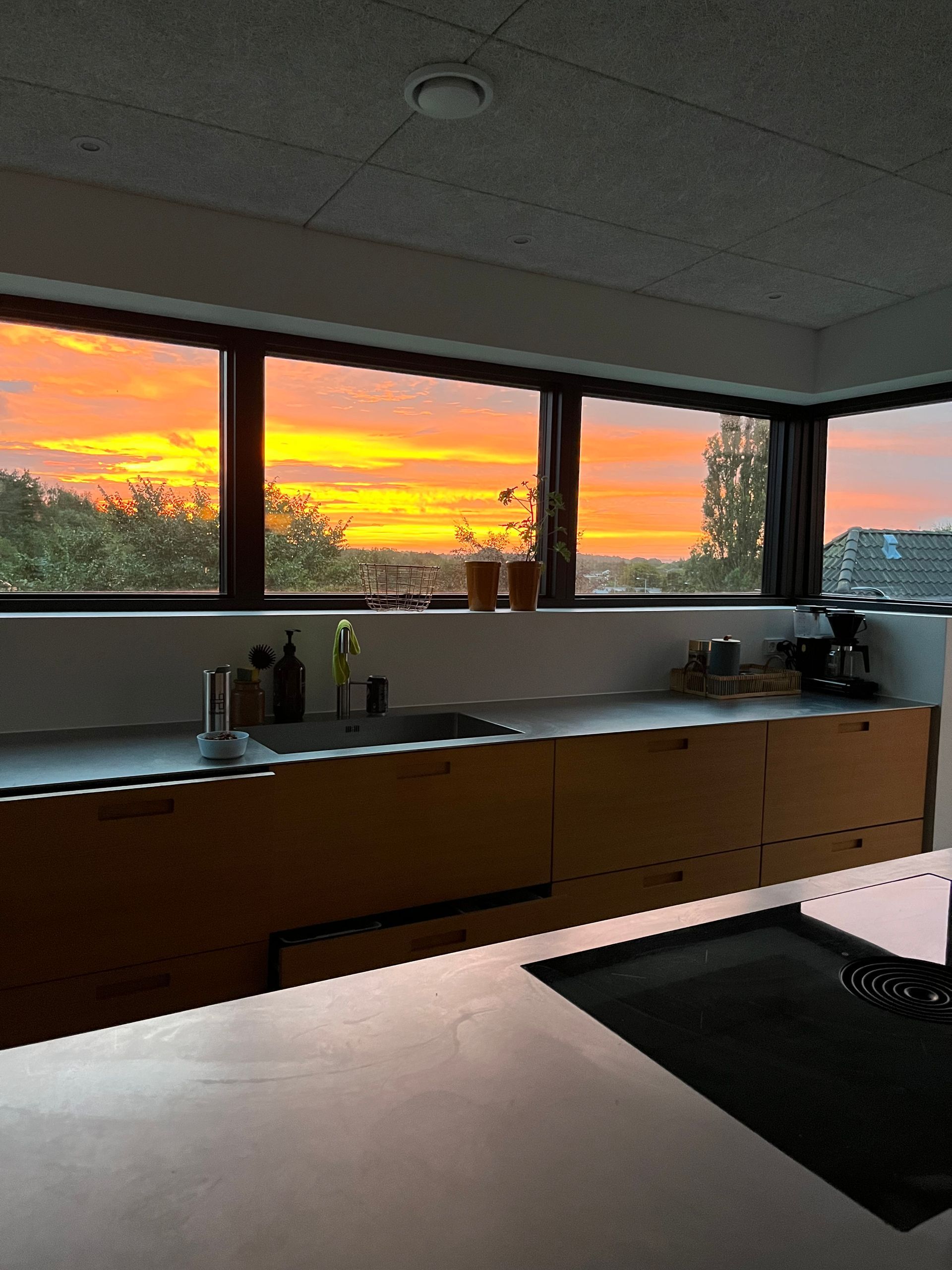Solnedgang over en køkkenbordplade og vask, med orange lys gennem store vinduer.