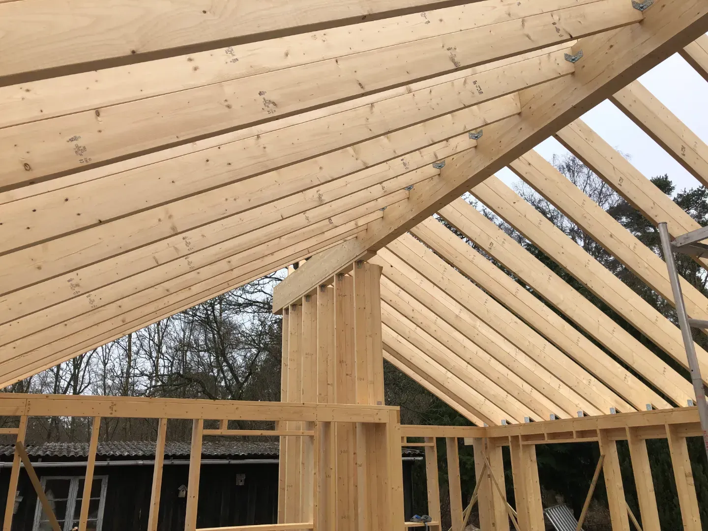 Træramme af et hus under opførelse, blå himmel, byggematerialer og et delvist færdiggjort hus i baggrunden.