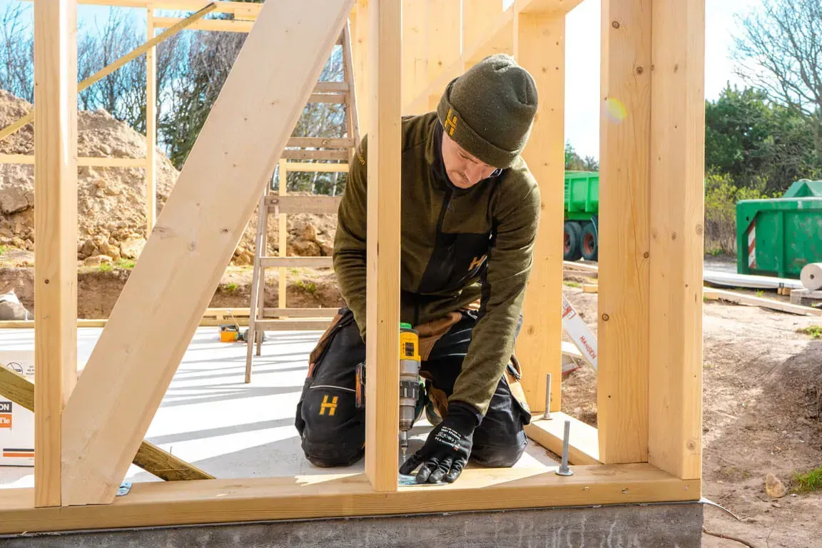 Træramme af et hus under opførelse, blå himmel, byggematerialer og et delvist færdiggjort hus i baggrunden.