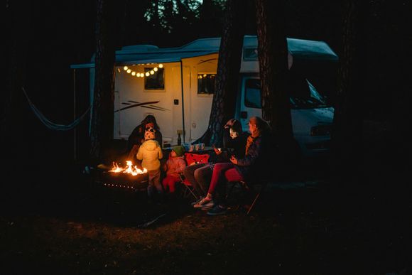 Family around campfire at night, RV in background, string lights, dark forest setting.