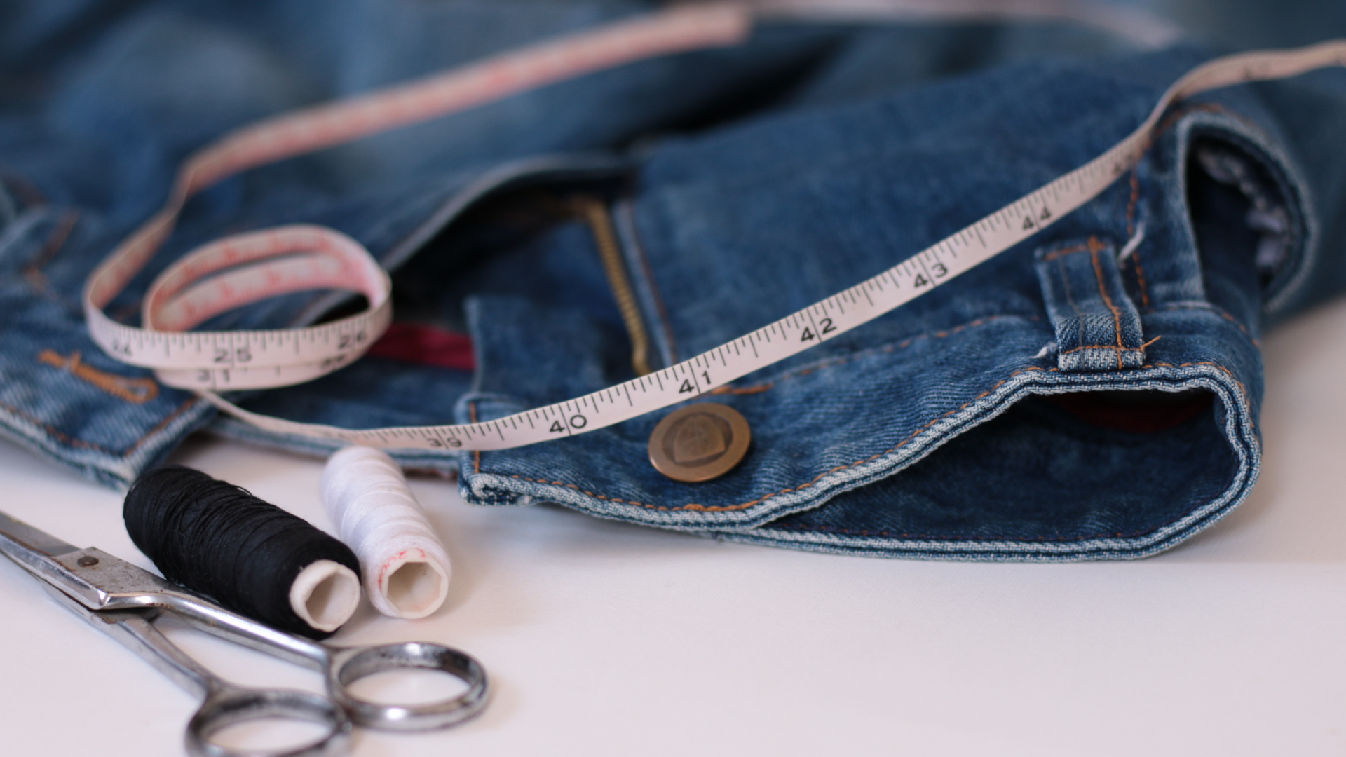 Denim jeans with measuring tape, thread spools, and scissors on a white surface