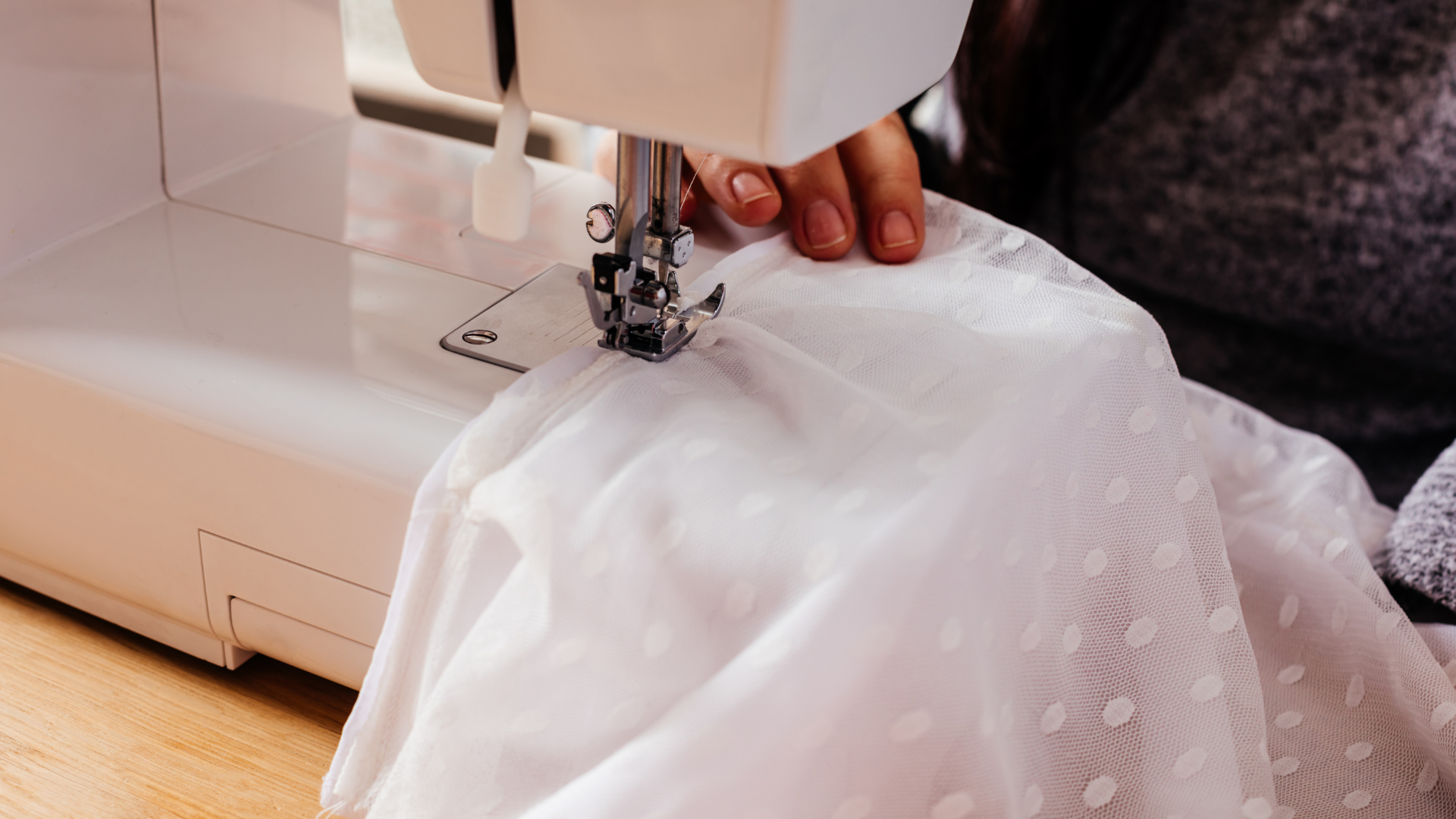 Hands guiding white fabric through a sewing machine, stitching a sheer garment.