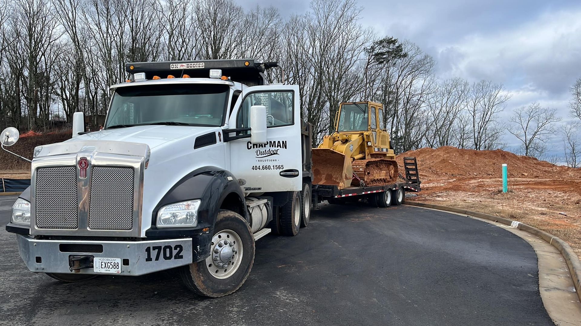 Two dump trucks with raised beds at a gravel site, one red and one white, with trees in the background.
