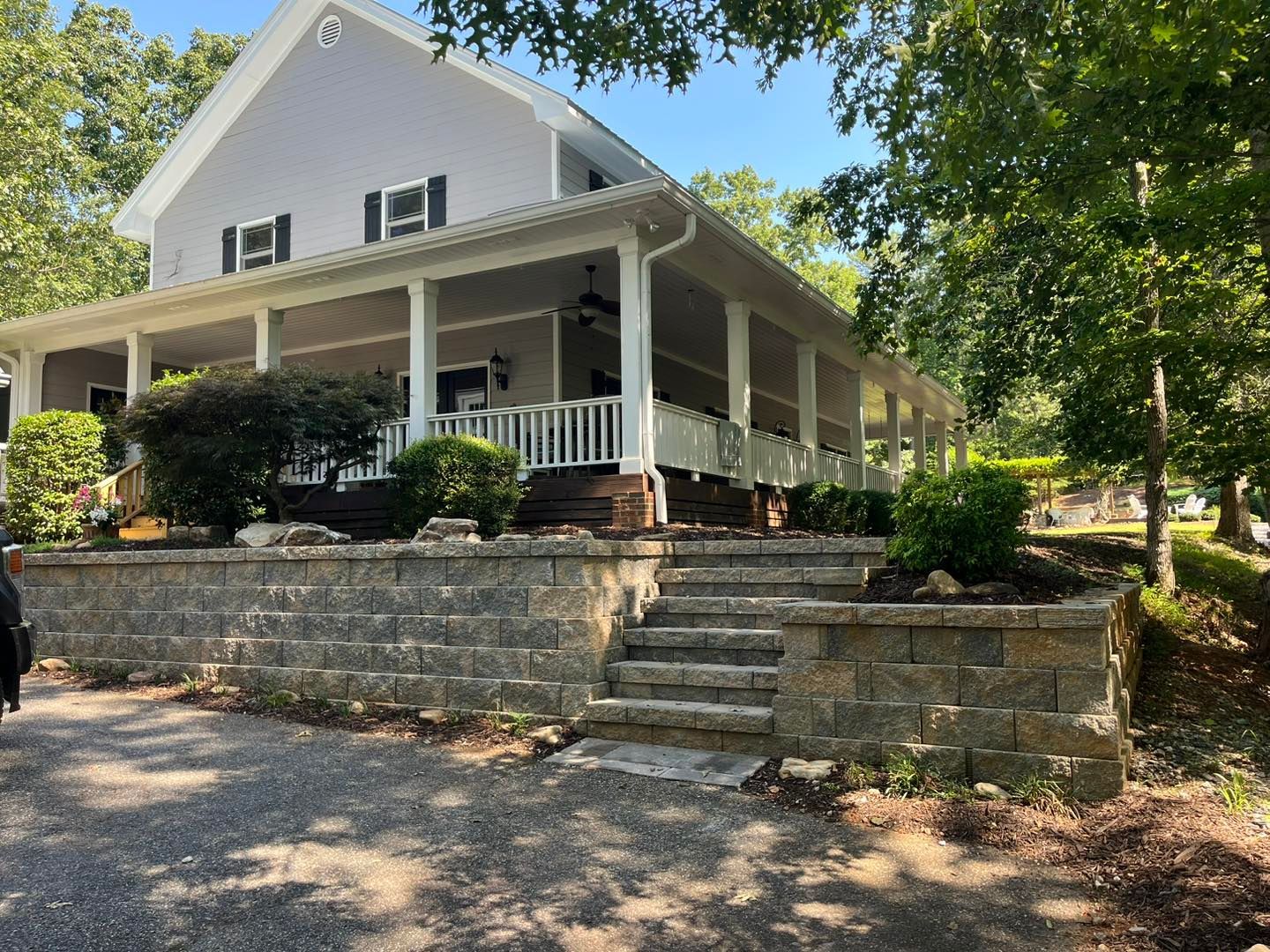 White farmhouse with wraparound porch on a stone retaining wall, surrounded by trees and gravel driveway