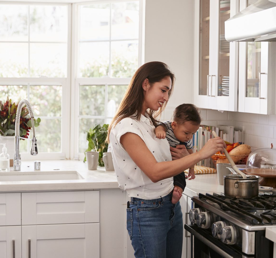 Woman in kitchen stirring pot while holding baby.
