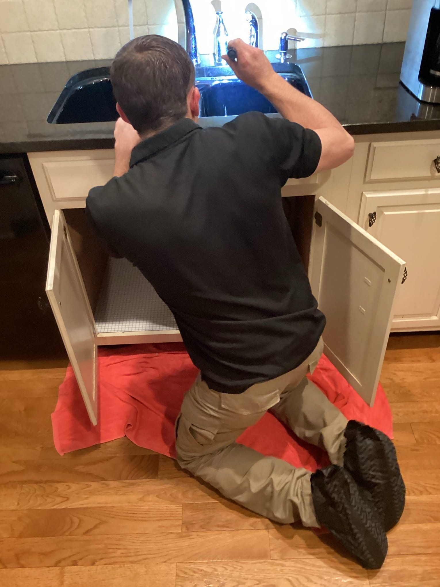 Person kneeling on kitchen floor repairing an open sink cabinet under a countertop