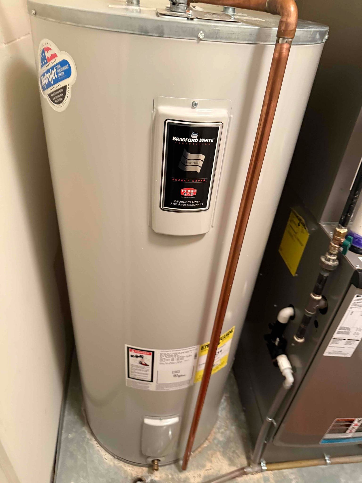 Water heater in a utility room; tall, silver tank with copper piping and control panel.