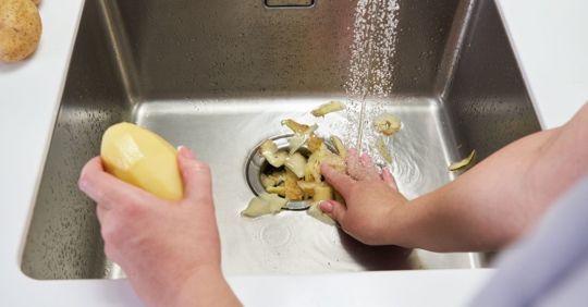 Hands washing potato peelings down a kitchen sink drain.