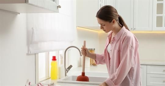 Woman using a plunger in a white kitchen sink, looking down.