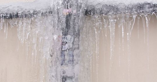 Ice-covered pipes against a beige wall. Icicles hang down from the frozen structure.