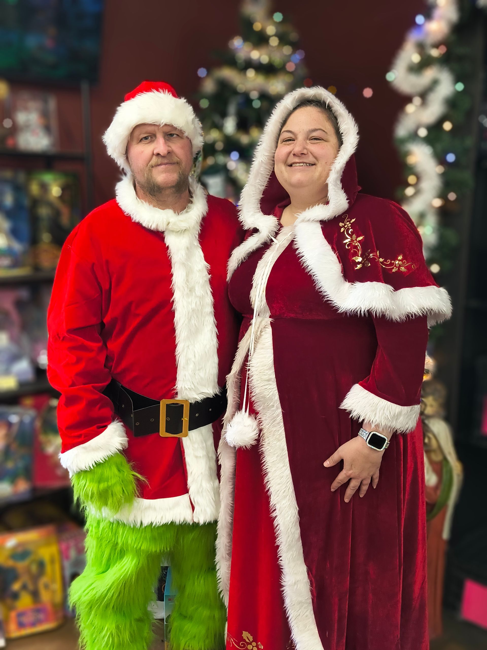 Two people in red and white Santa outfits posing in front of a decorated Christmas tree.