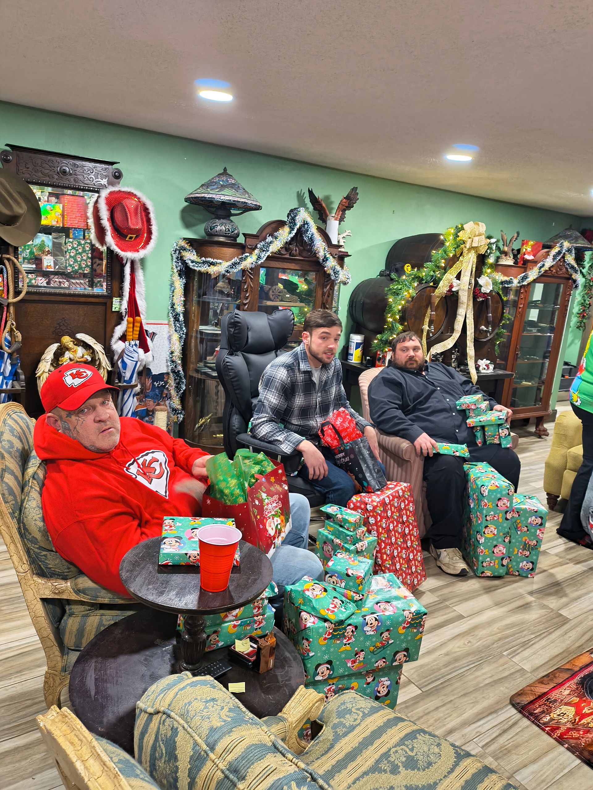People seated among wrapped gifts in a festive holiday room with decorated trees and ornaments