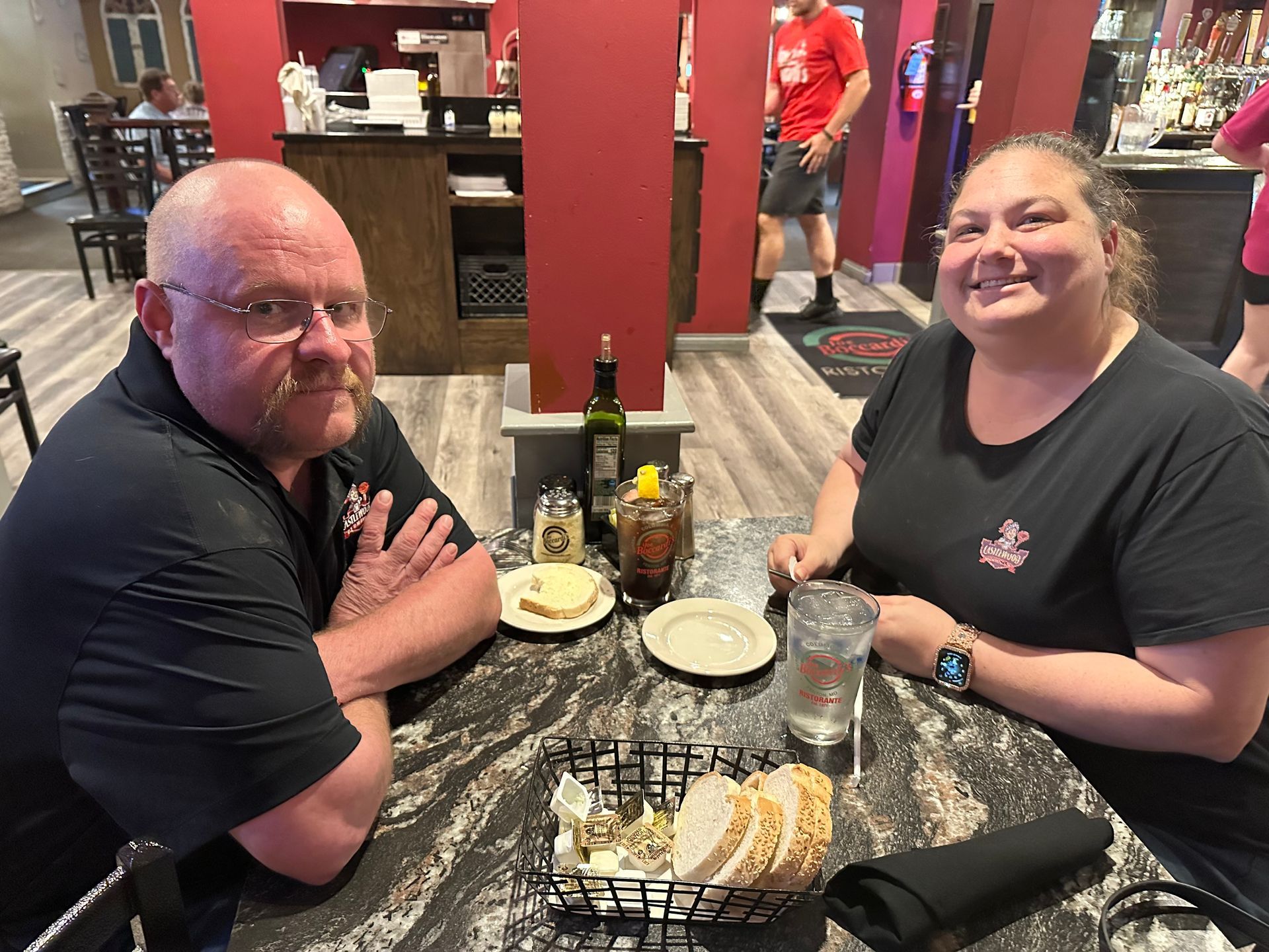 Two people sitting at a restaurant table with food. Man on left, woman on right, smiling, inside a dining area.
