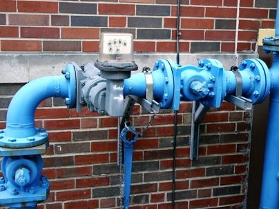 Blue industrial water pipe with various valves and a meter against a brick wall.