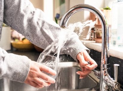 Person filling a glass with water at a sink, water splashing on the person's sleeve.