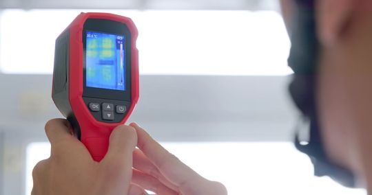 Person using a red and black thermal imaging camera, examining a wall.
