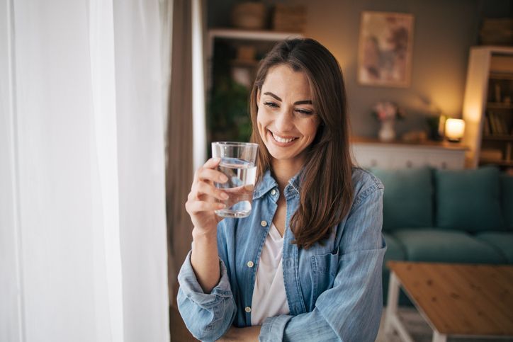 Woman smiling, holding a glass of water. She's in a cozy living room with a blue couch and natural light.