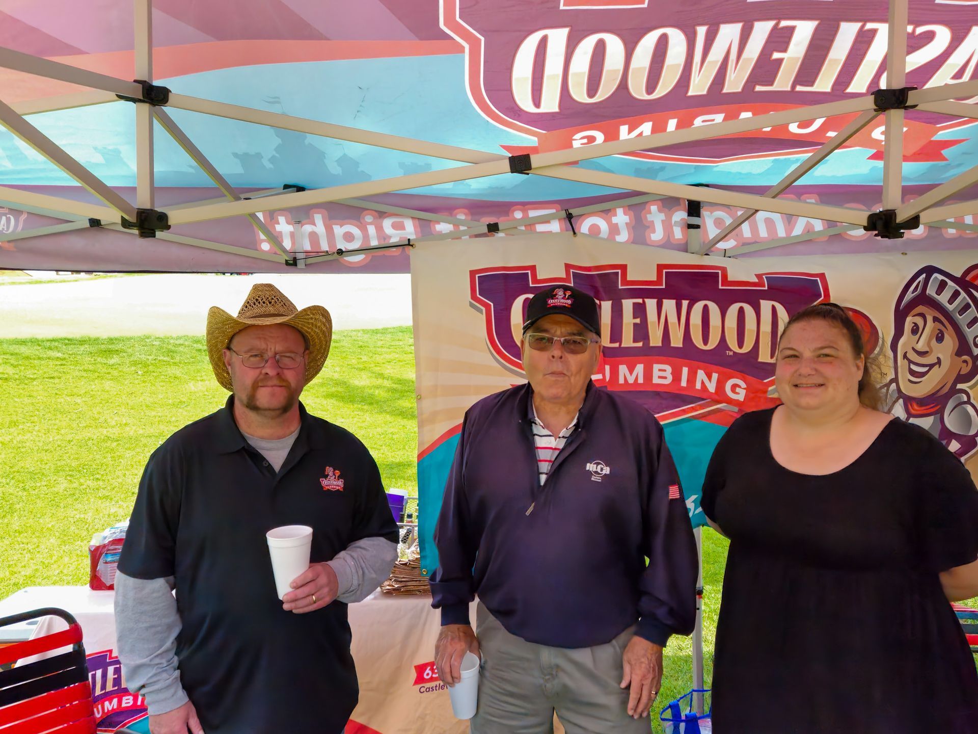 Three people stand at an outdoor tent for Castlewood Plumbing. Two men and a woman smile. A man in a cowboy hat holds a cup.
