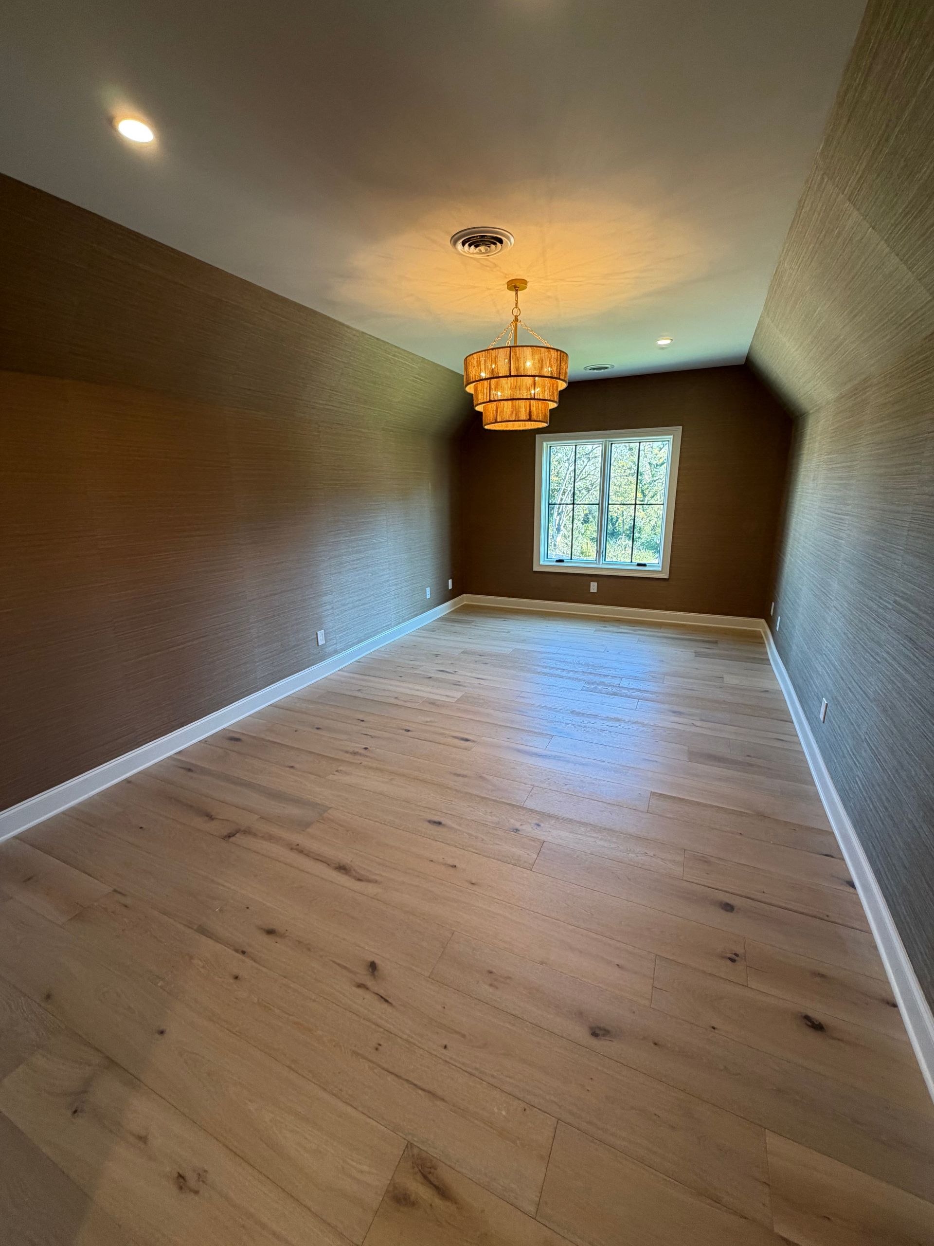 Empty long room with light wood floors, textured brown walls, chandelier, and a window at the far end.