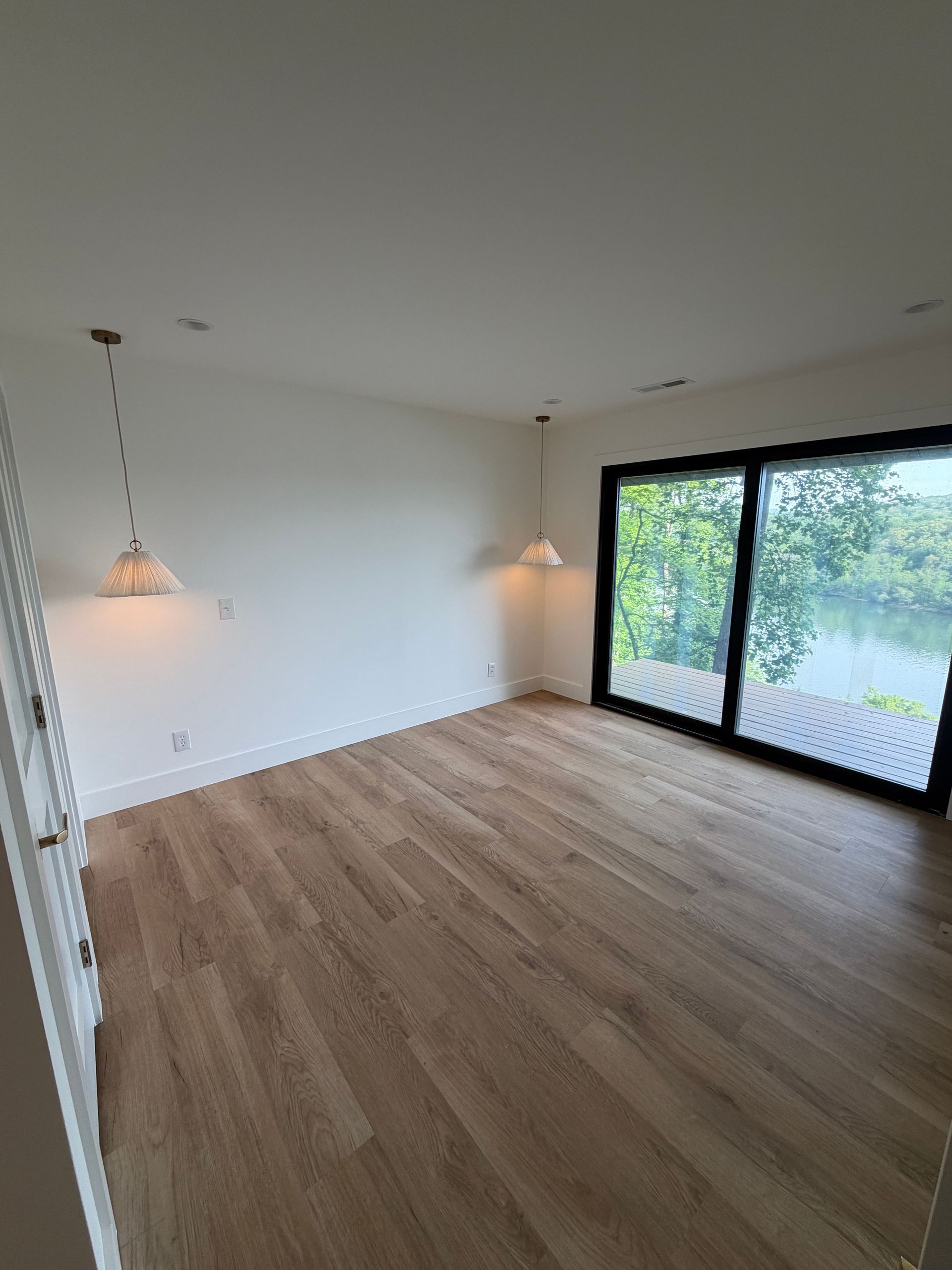 Empty modern living room with wood floors, white walls, pendant lights, and large sliding glass doors to a garden.