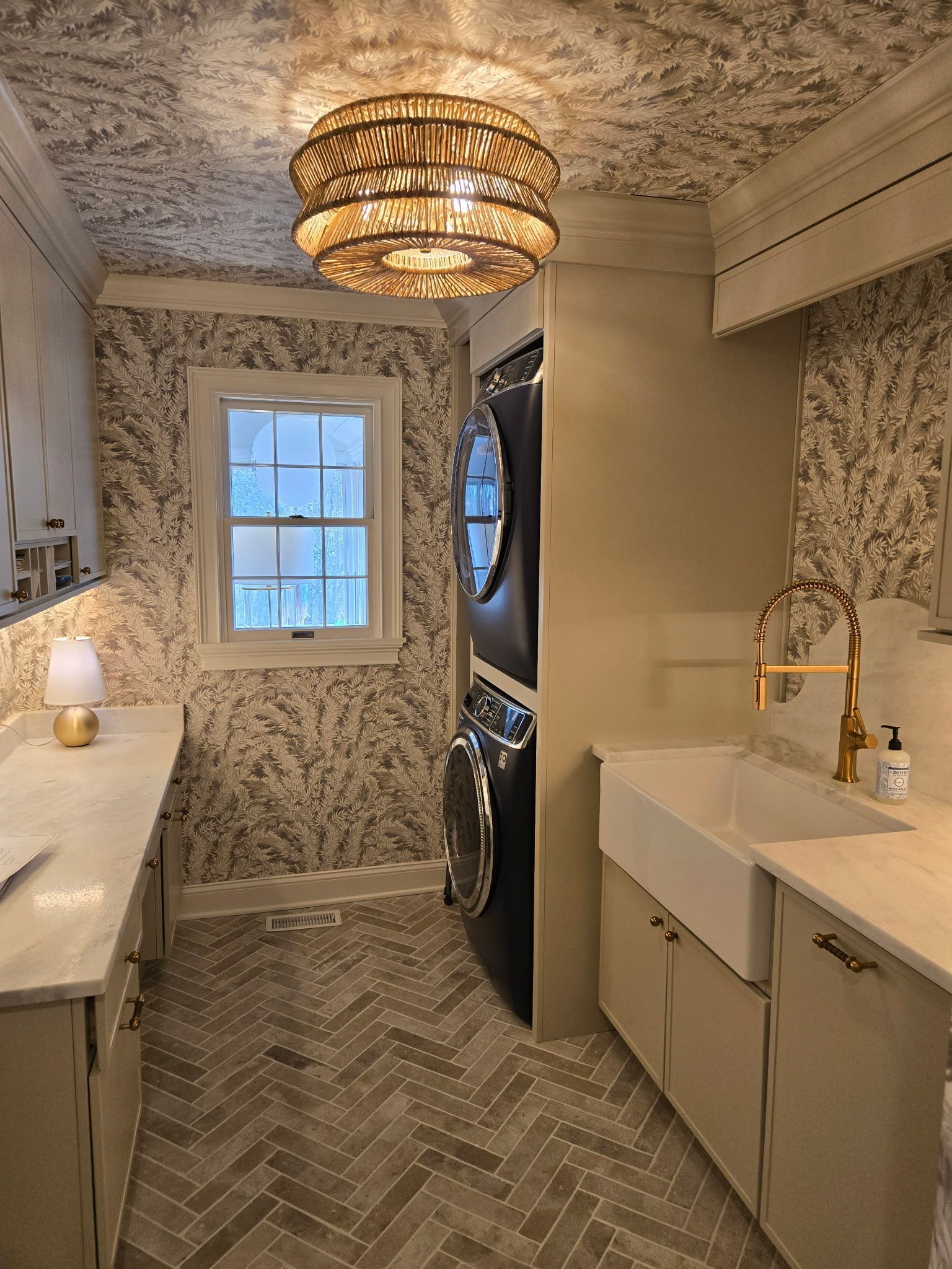 Narrow laundry room with patterned wallpaper, stacked washer and dryer, utility sink, window, and woven ceiling light