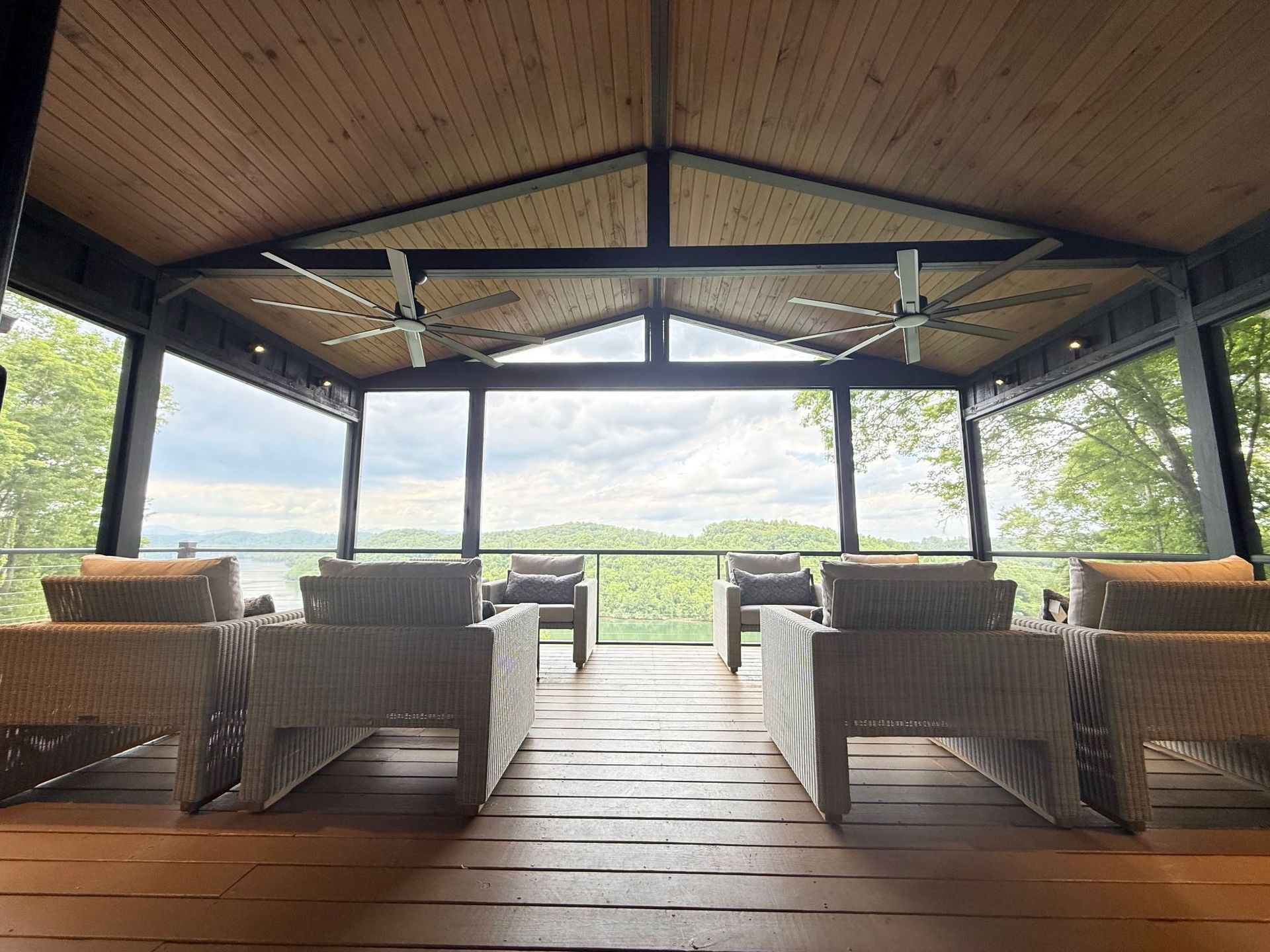 Covered patio with wicker chairs and wooden deck, overlooking a green landscape through open sides.