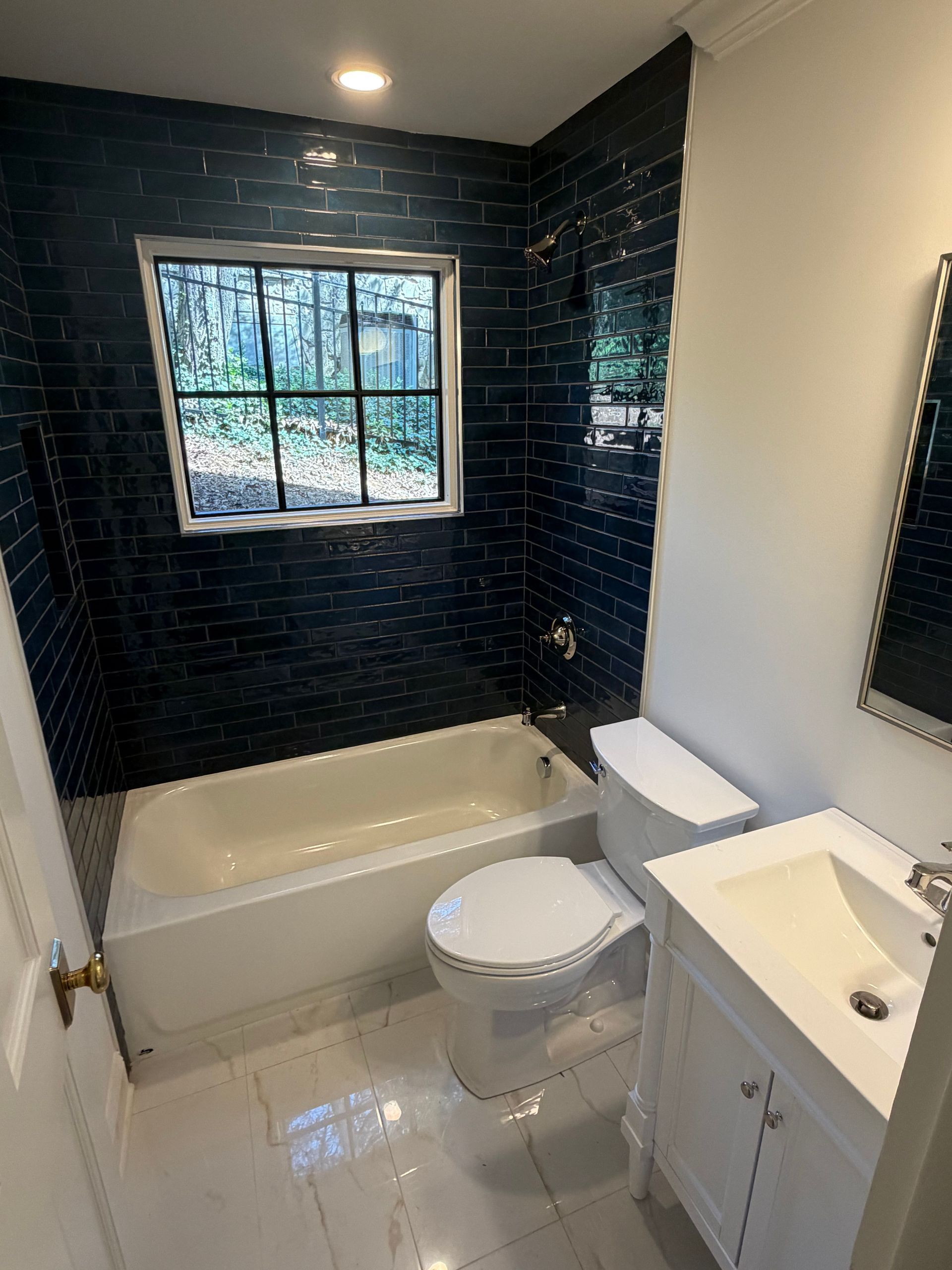 Small bathroom with black tile shower, white tub, toilet, and vanity under a window.