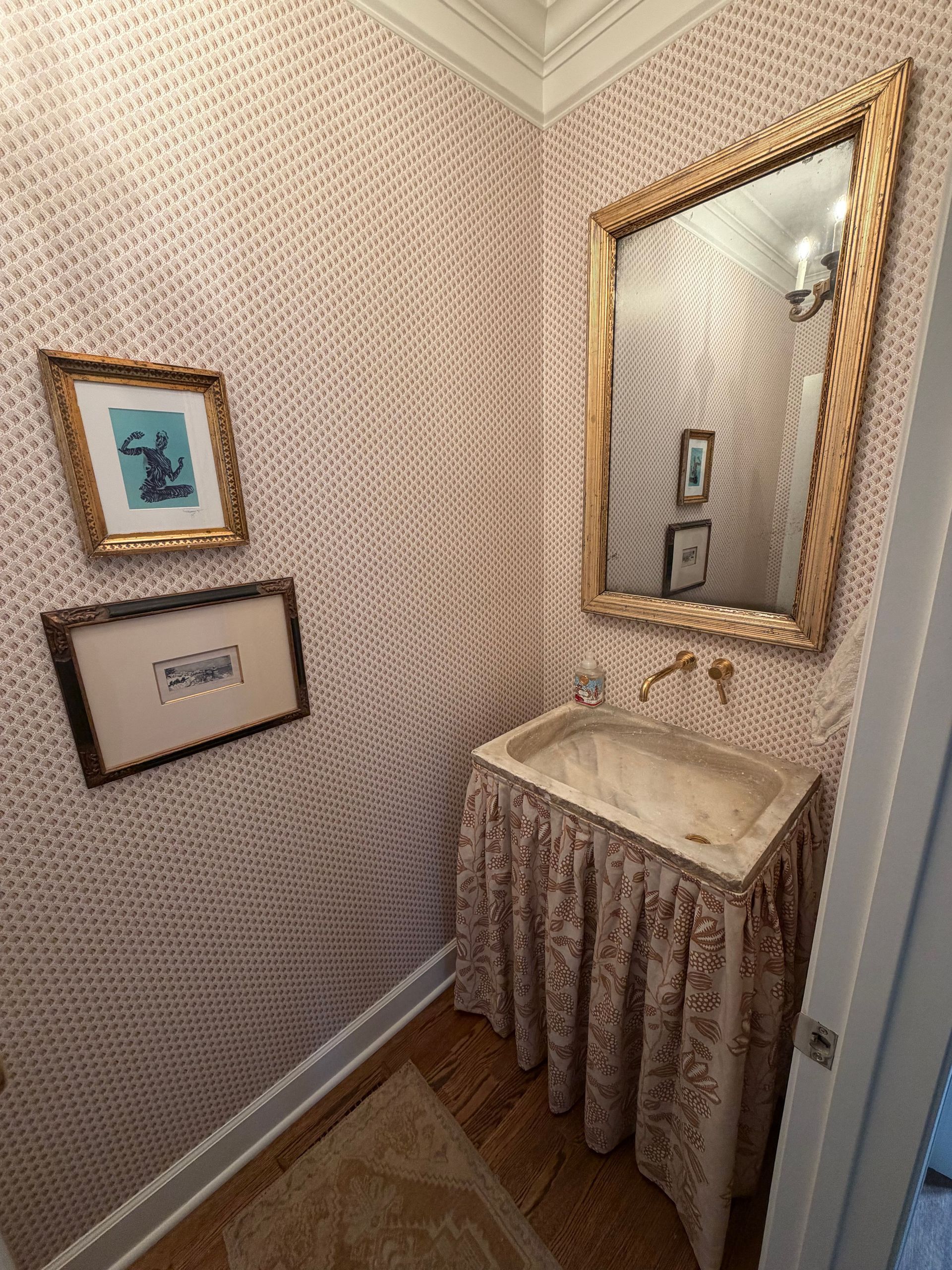 Small powder room with patterned wallpaper, pedestal sink, gold-framed mirror, and framed wall art.