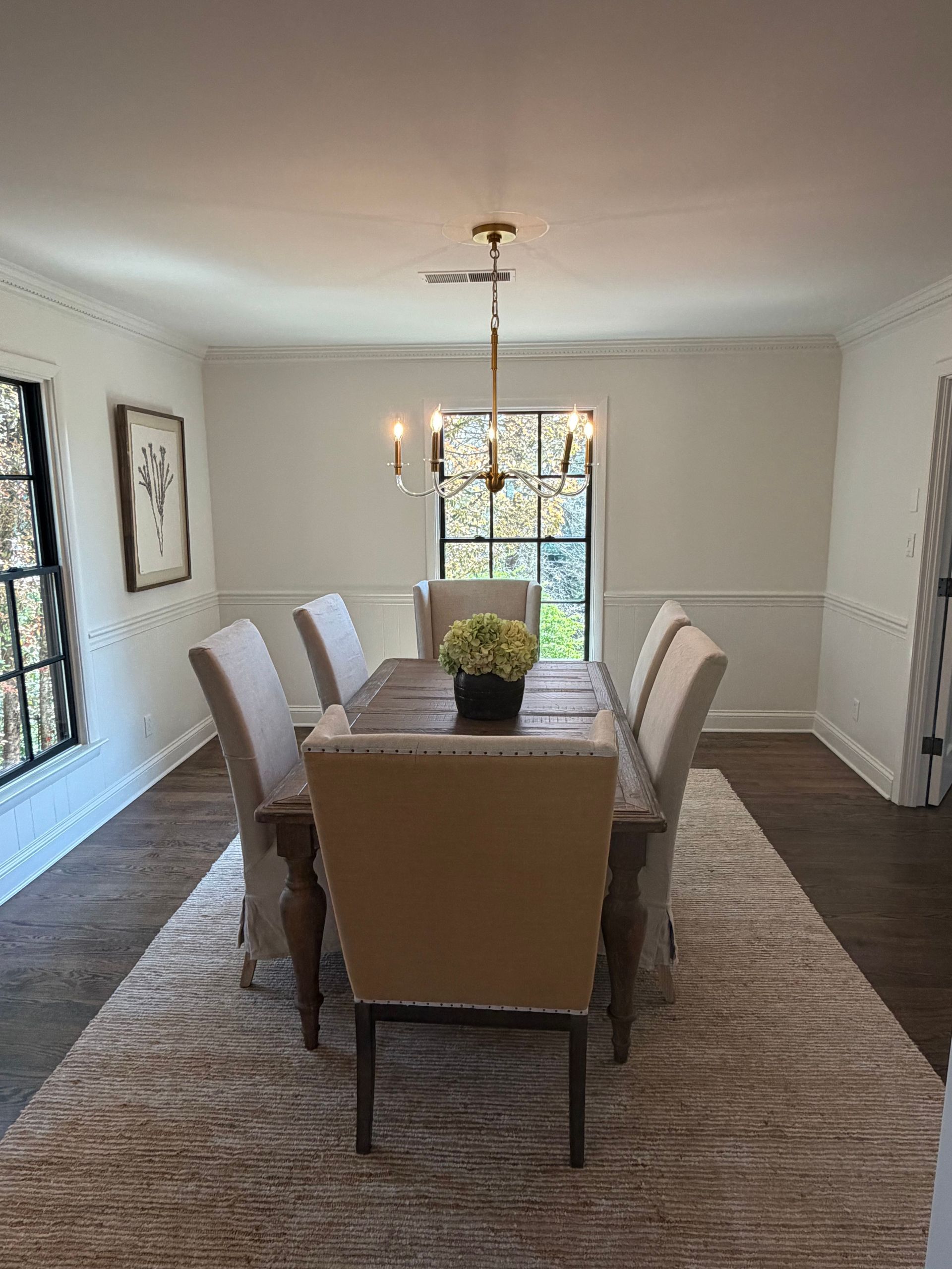 Dining room with long table, upholstered chairs, rug, and chandelier by bright windows