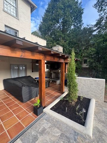 Covered patio with terracotta tiles, wooden pergola, black sofa, and planter beside a house.